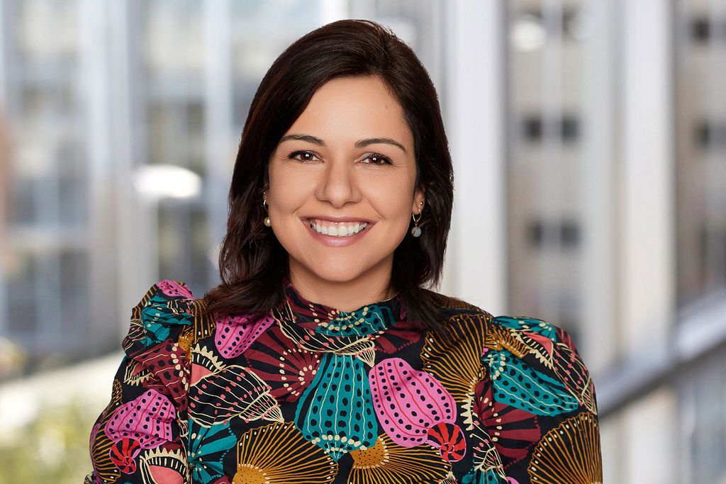 A headshot of Vivi Caleffi-Prichard smiling warmly and wearing a colorful blouse features a bold, abstract floral or sea-inspired pattern with shades of teal, pink, yellow, red, and black against softly blurred modern office buildings in the background